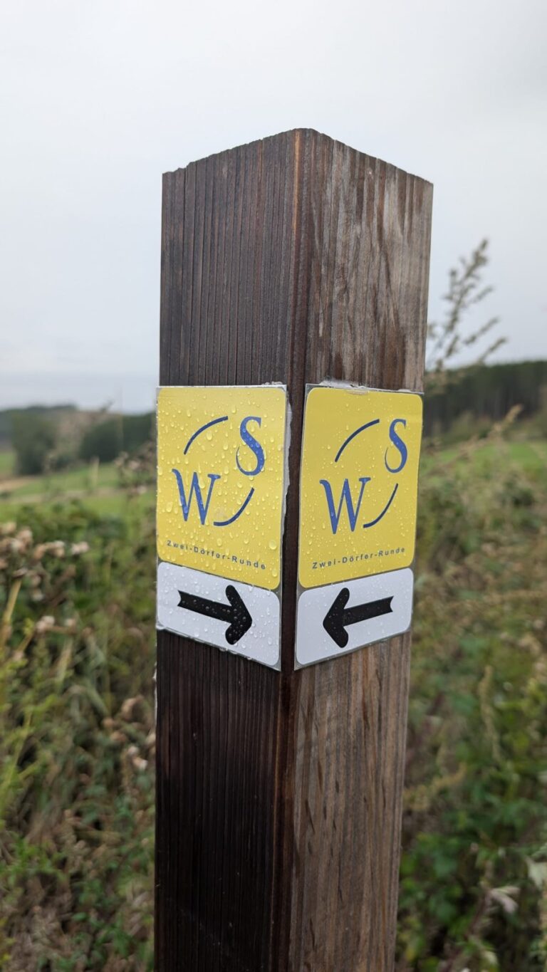 Holzpfosten mit gelben Wegweisern „WS – Zwei-Dörfer-Runde“ und Pfeilen